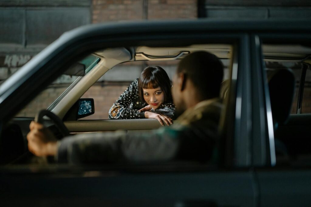 A woman in a stylish outfit leans on a car window while engaging with a driver inside.