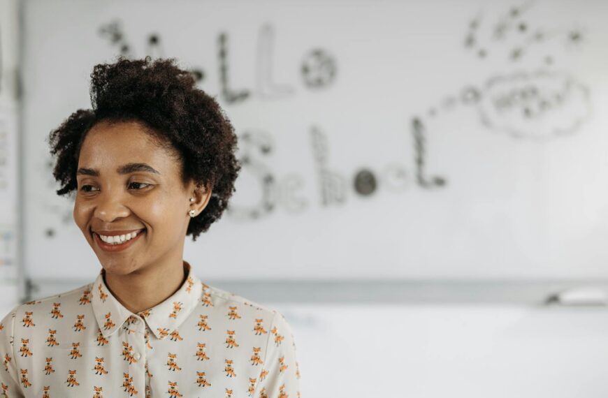 A friendly female teacher smiles in front of a classroom whiteboard.