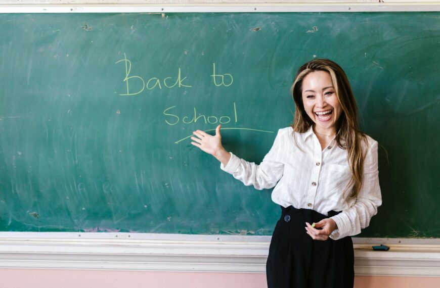 Smiling teacher in a classroom pointing at back to school message on a chalkboard.