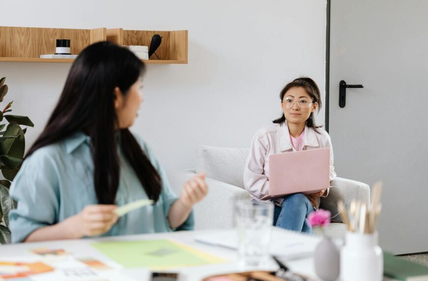 Two women engage in a discussion in a modern office setting with laptops.