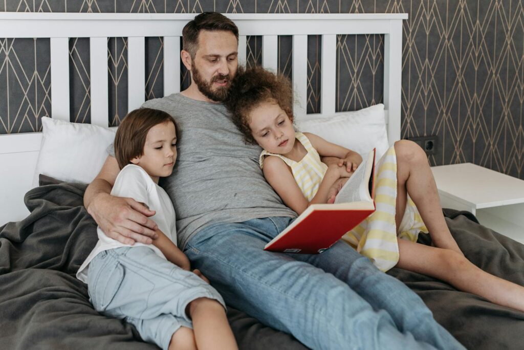 A father reads a storybook to his two children, creating a warm family moment in a cozy bedroom.
