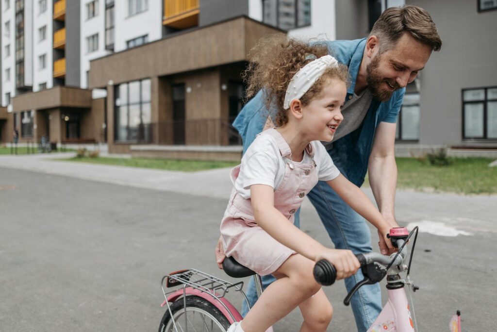 Father helps daughter learn to ride a bike in an urban setting, capturing joy and bonding.