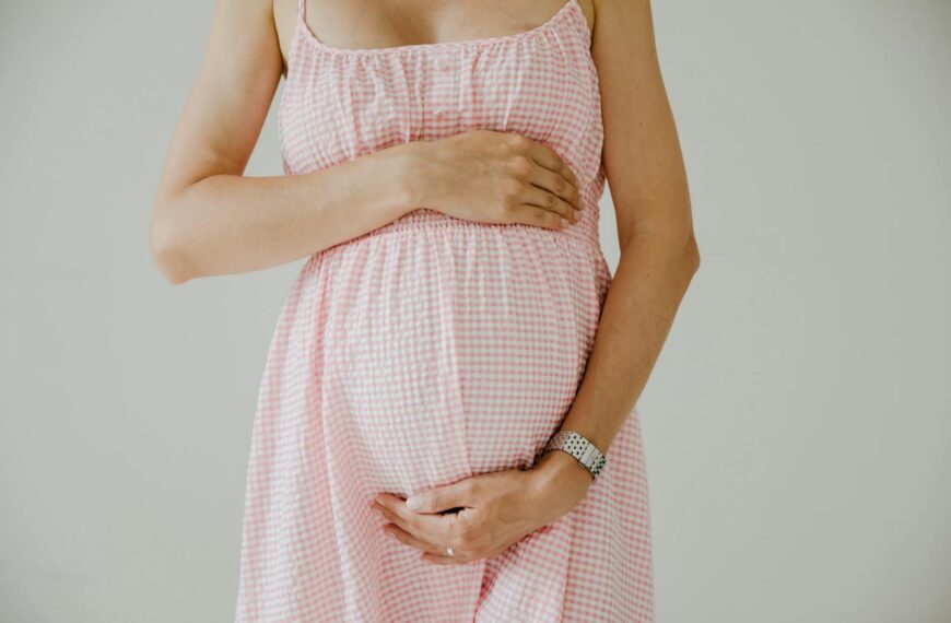 Close-up of pregnant woman holding her belly, wearing a pink checkered dress indoors.