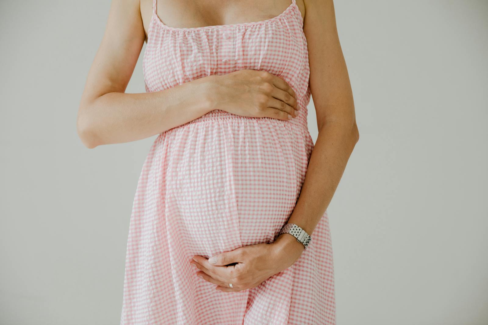 Close-up of pregnant woman holding her belly, wearing a pink checkered dress indoors.