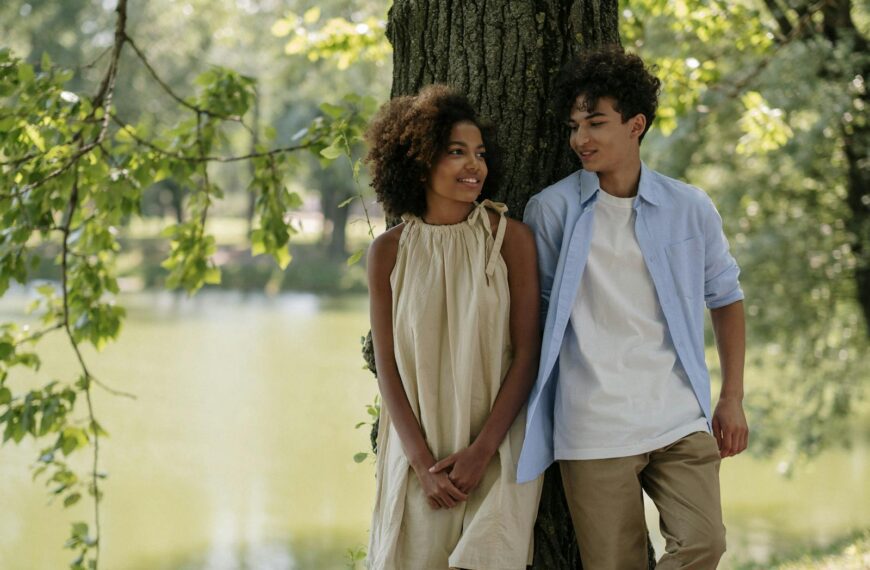 A young couple with afro hair enjoying a sunny day by the lake, smiling and leaning against a tree.