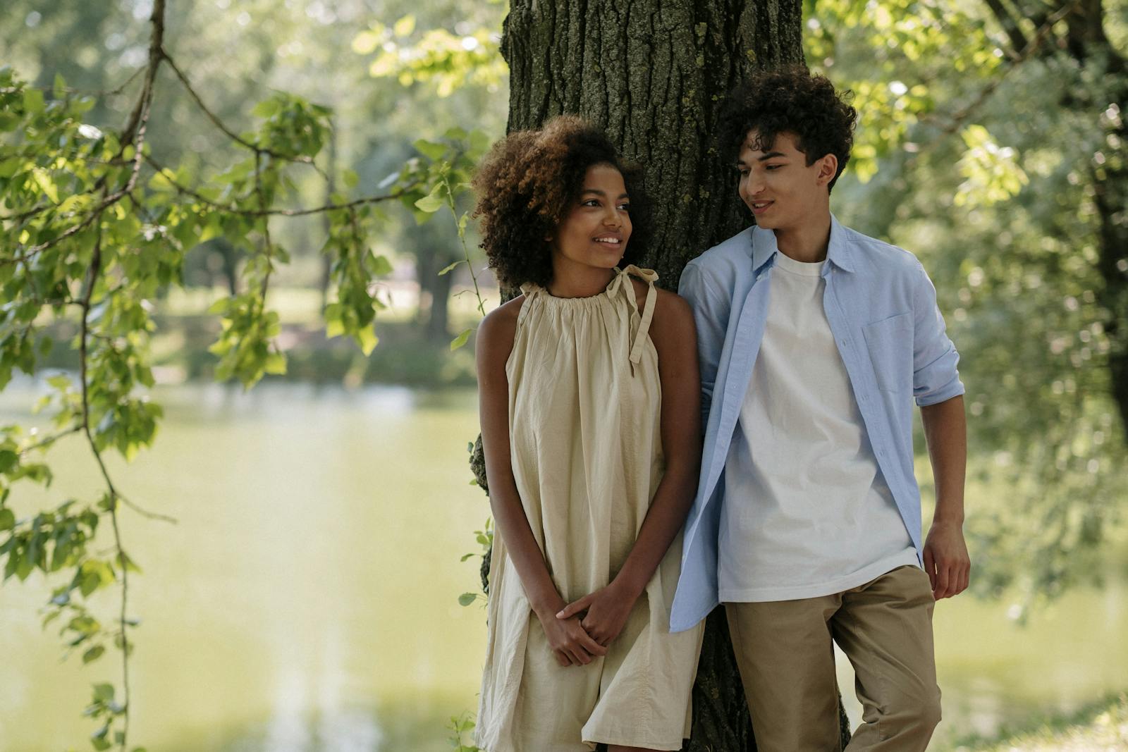 A young couple with afro hair enjoying a sunny day by the lake, smiling and leaning against a tree.