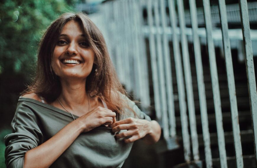 Smiling woman in an outdoor setting, wearing a casual gray shirt near a fence.