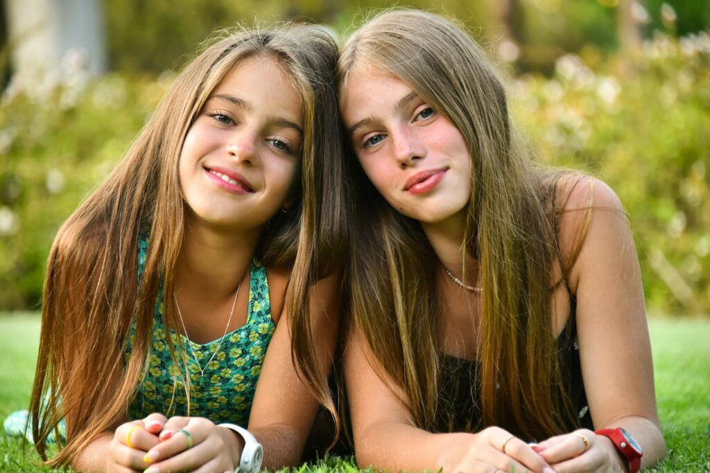 Two smiling teenage girls lying on grass in a sunny outdoor setting, embracing summer vibes.