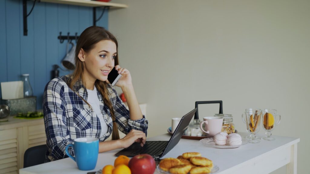 Woman talking on phone while working on laptop at table.