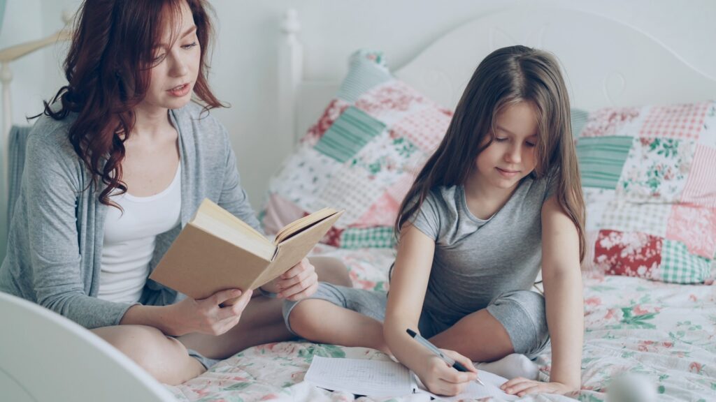 Mom reads while daughter draws on the bed.