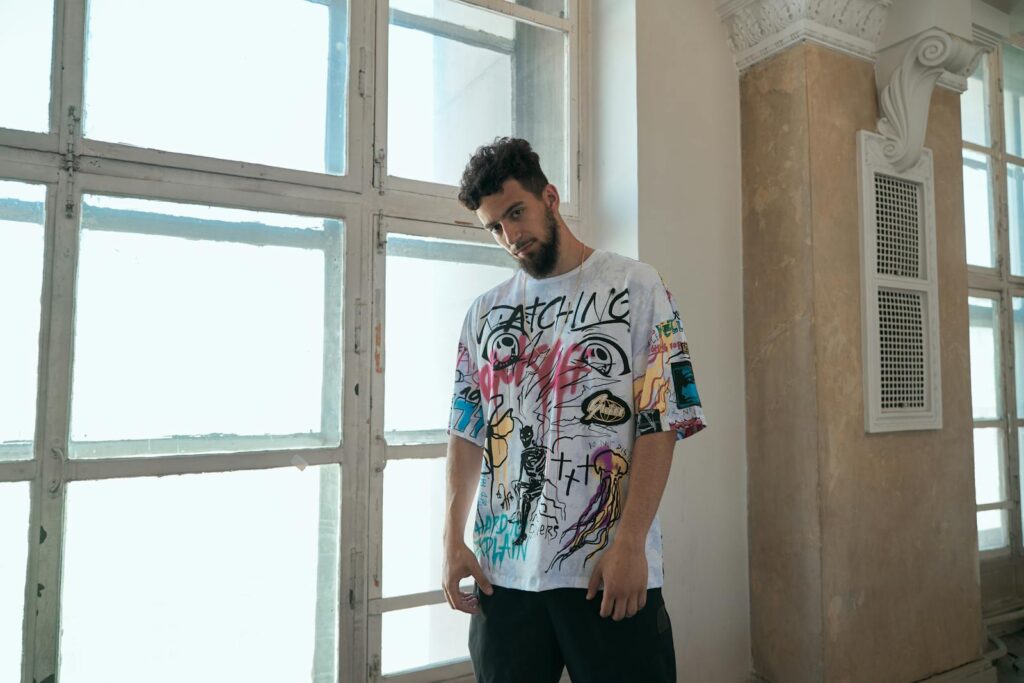 Man in a graffiti t-shirt posing stylishly indoors with natural window light.