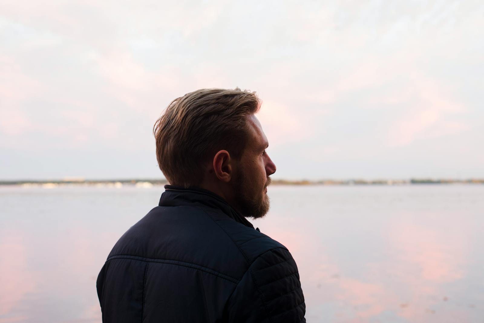 A serene side profile shot of a man gazing over a calm lake at sunset, capturing a moment of reflection.