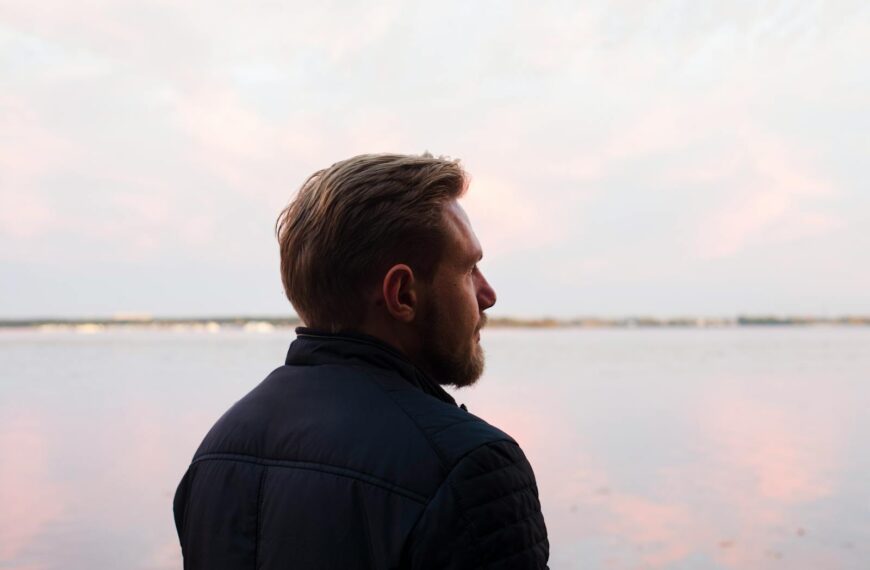 A serene side profile shot of a man gazing over a calm lake at sunset, capturing a moment of reflection.
