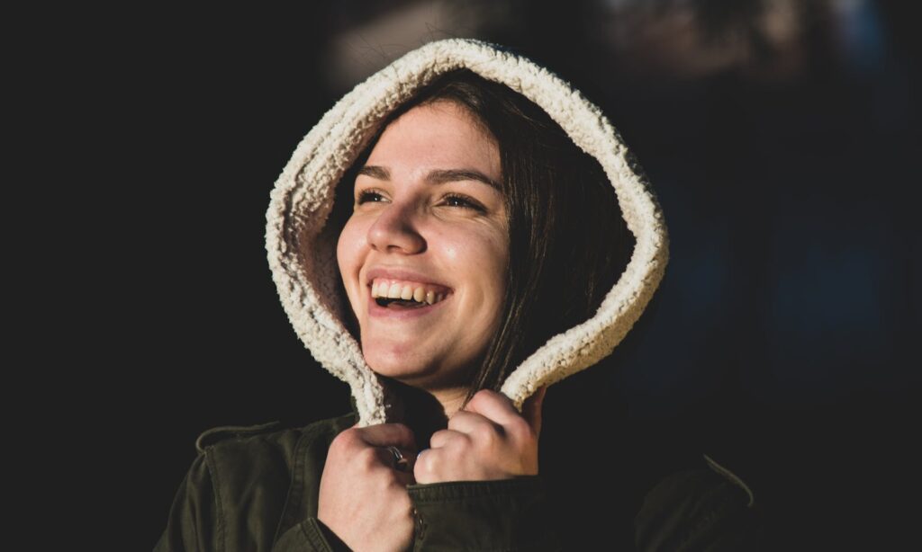 Cheerful woman smiling warmly with a hooded jacket against a dark background.