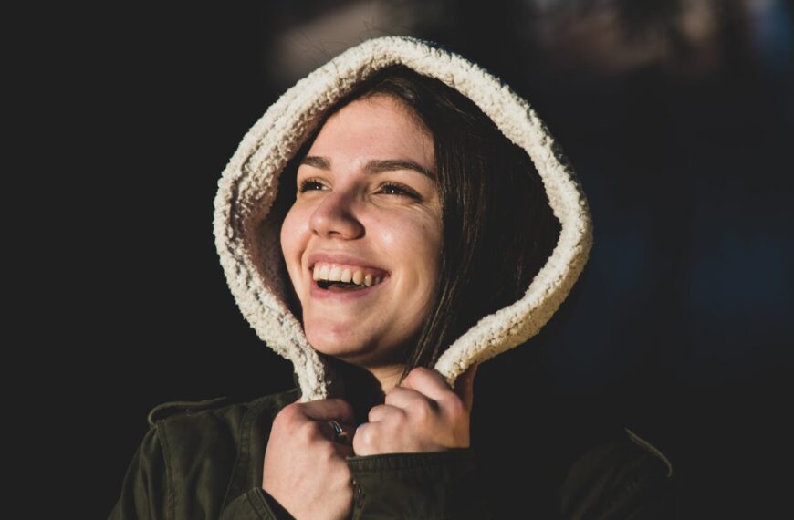 Cheerful woman smiling warmly with a hooded jacket against a dark background.