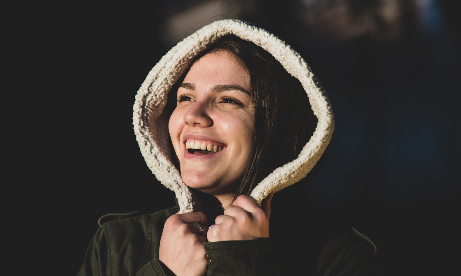 Cheerful woman smiling warmly with a hooded jacket against a dark background.