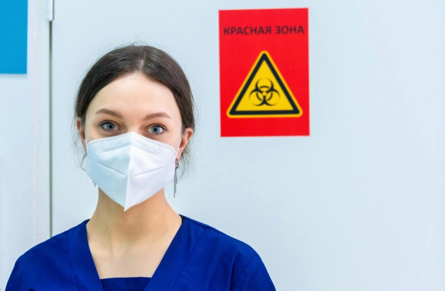 Healthcare professional in protective gear standing near a biohazard warning sign.