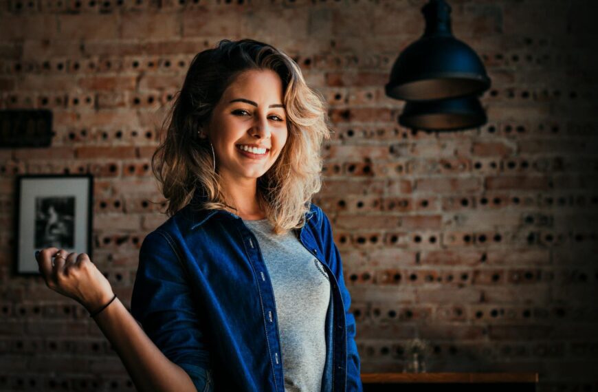 Portrait of a smiling woman in denim jacket against a brick wall indoors.