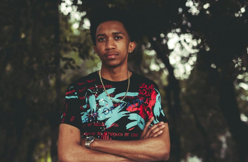 Stylish young man with afro hair stands confidently outdoors with arms crossed, wearing a graphic t-shirt.