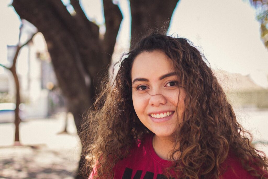 Portrait of a smiling young woman with curly hair in a sunny outdoor setting.