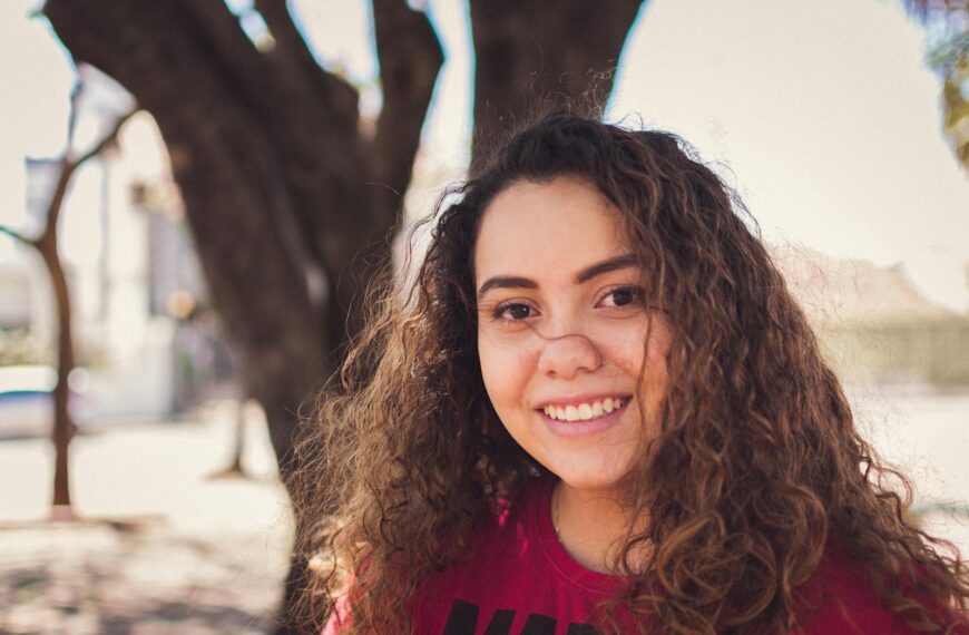 Portrait of a smiling young woman with curly hair in a sunny outdoor setting.