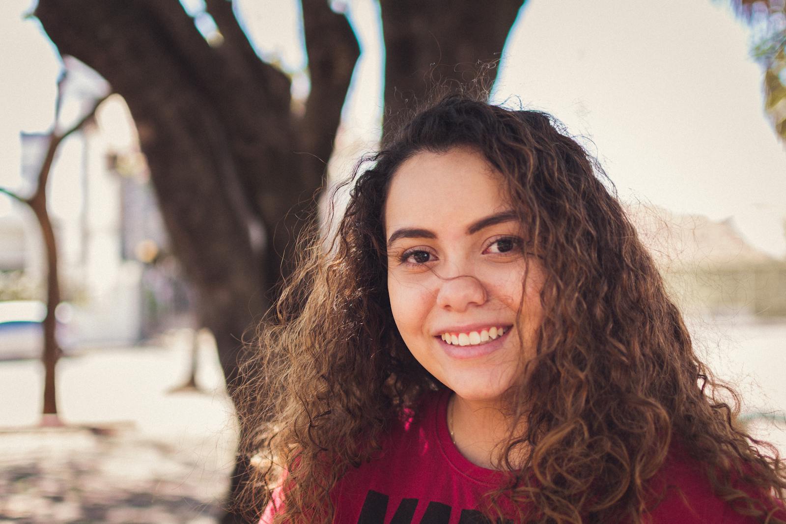 Portrait of a smiling young woman with curly hair in a sunny outdoor setting.
