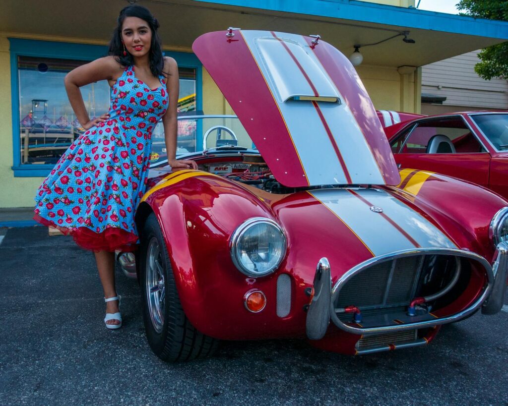 A retro-style woman in a floral dress stands beside a classic red car with the hood open.