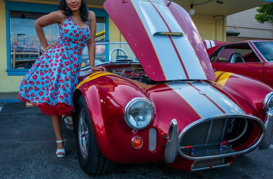 A retro-style woman in a floral dress stands beside a classic red car with the hood open.