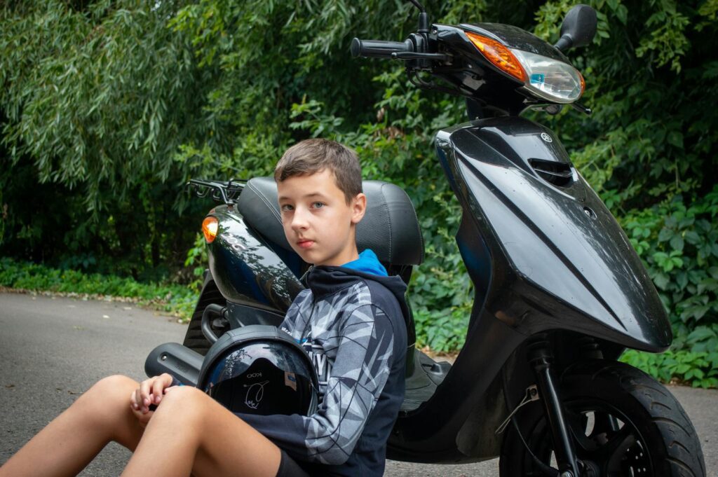 A young boy sitting outdoors with a black scooter and helmet, surrounded by lush greenery.