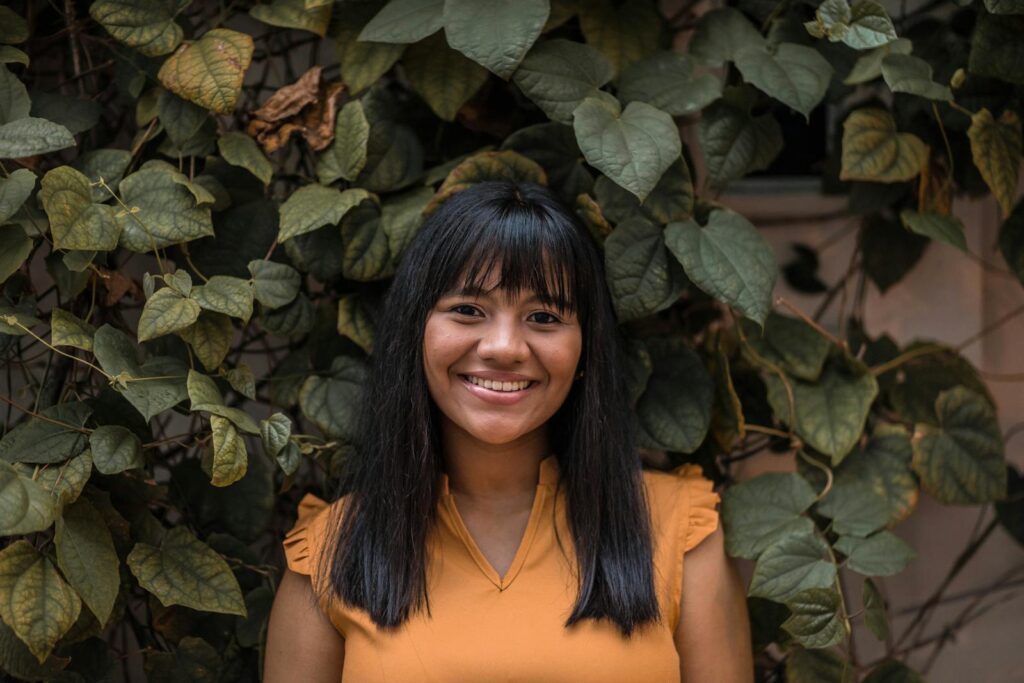 A cheerful woman stands against a backdrop of lush green leaves, radiating positivity.