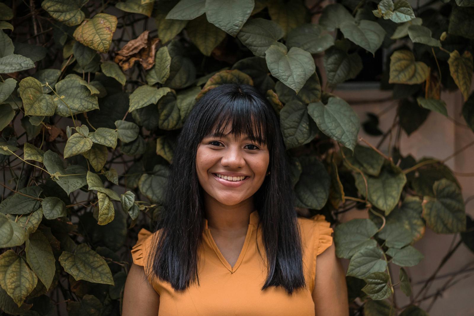 A cheerful woman stands against a backdrop of lush green leaves, radiating positivity.