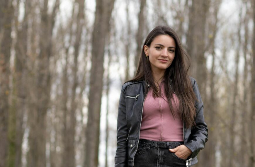 Portrait of a young woman with long hair wearing a black jacket, standing outdoors in a forest.