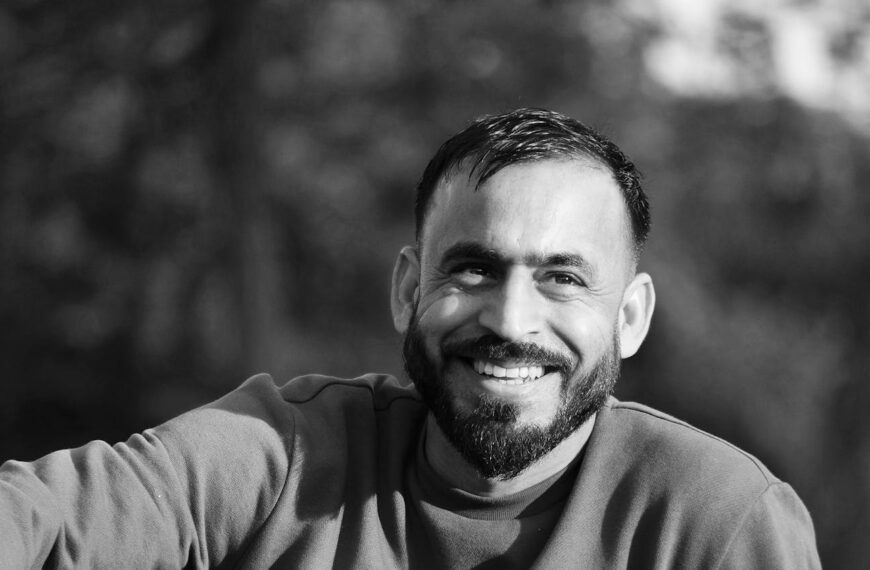 A black and white portrait of a smiling man with a beard in an outdoor setting.