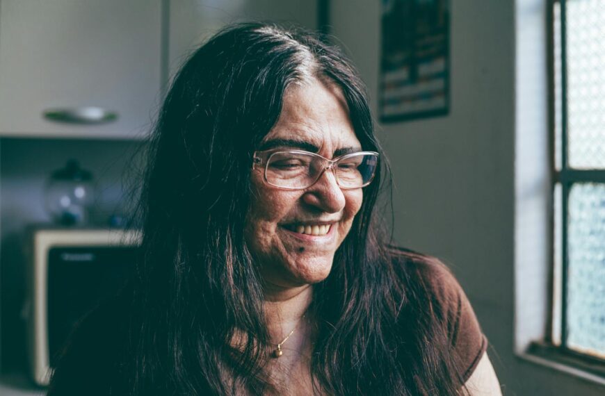 Elderly woman with glasses smiling in a kitchen setting.