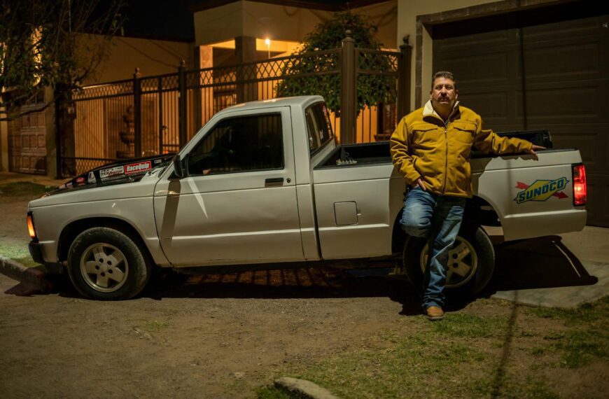 A man in a yellow jacket leaning on a white pickup truck at night in an urban area.