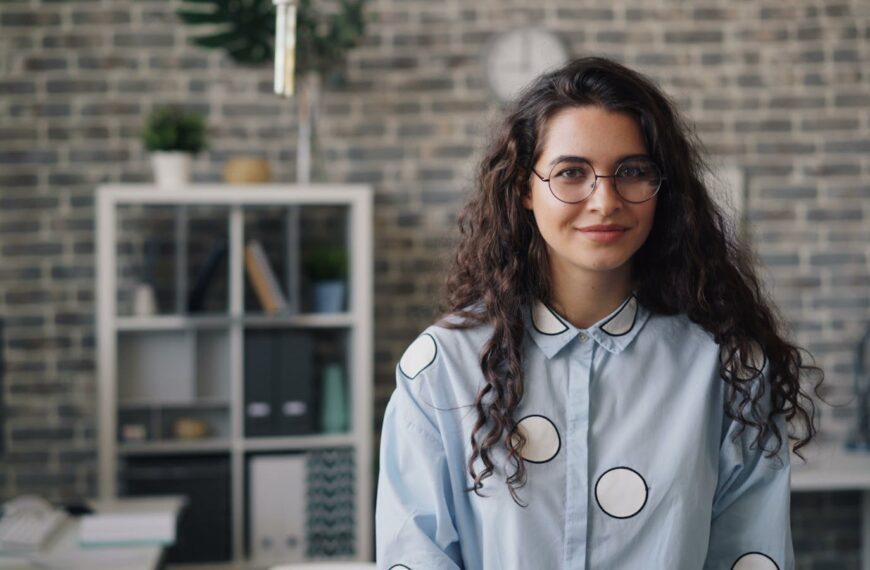 Smiling young woman in stylish office setting, wearing eyeglasses and patterned shirt.