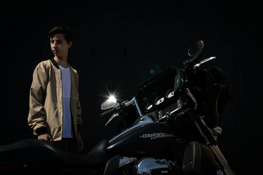 A young man in a brown jacket stands next to a parked motorcycle at night.