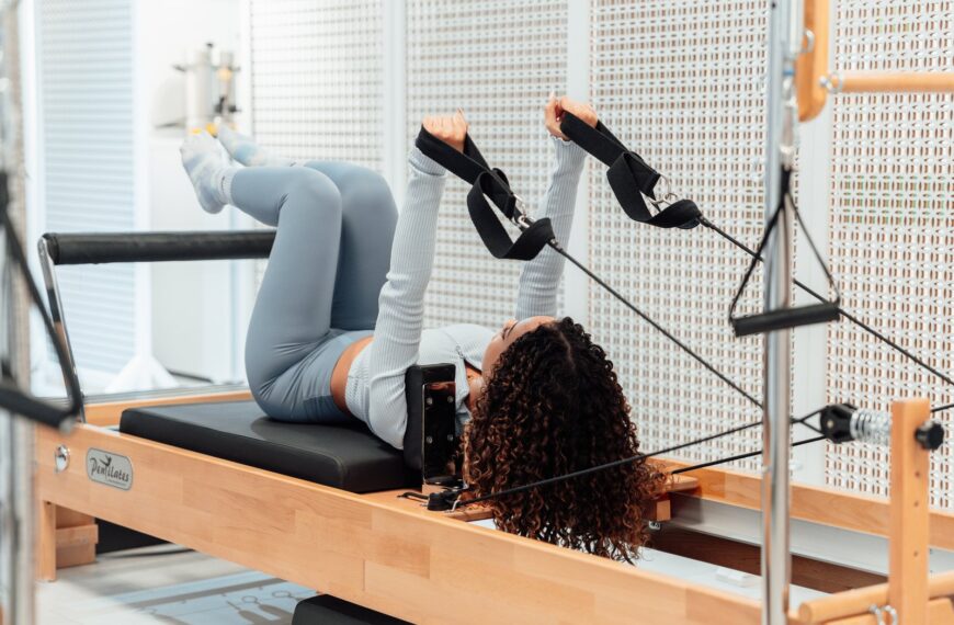 A woman wearing a gray tracksuit exercises on a Pilates reformer in a modern studio setting.