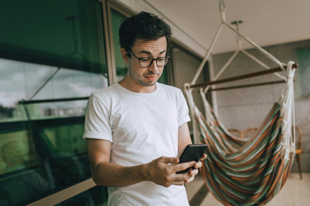 Young man in glasses using smartphone indoors with striped hammock.