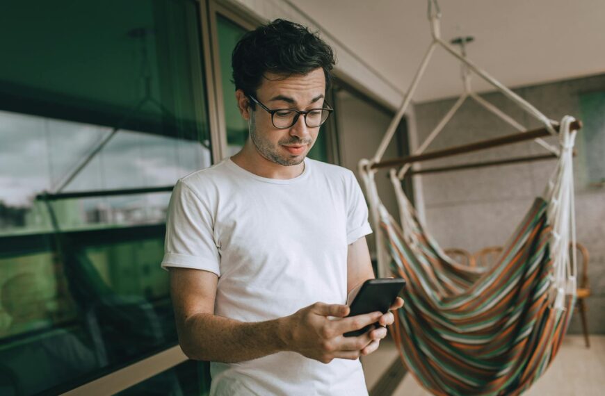 Young man in glasses using smartphone indoors with striped hammock.