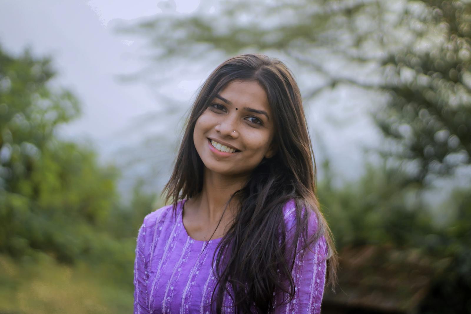 A young woman smiles warmly while standing outdoors, wearing a purple dress. Vibrant and natural setting.