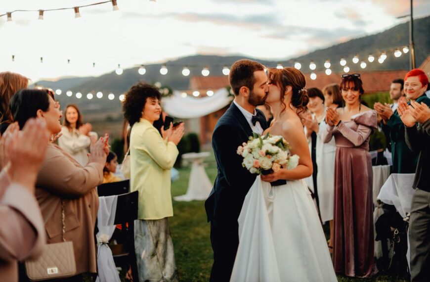 A bride and groom share a passionate kiss outdoors with guests applauding under string lights.