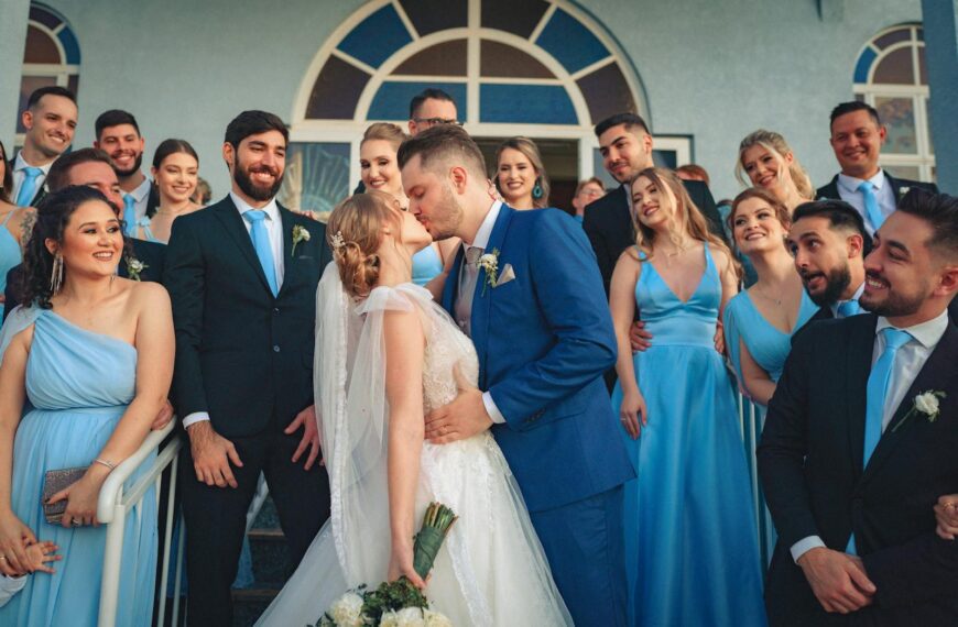 Bride and groom kiss surrounded by joyful wedding party in blue attire.