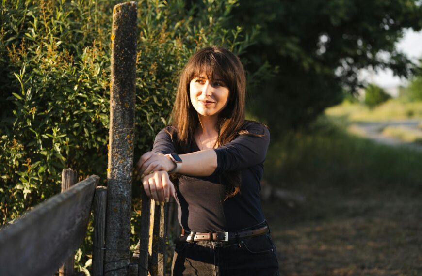A young woman leans on a fence in the countryside during a beautiful sunset, enjoying the peaceful atmosphere.