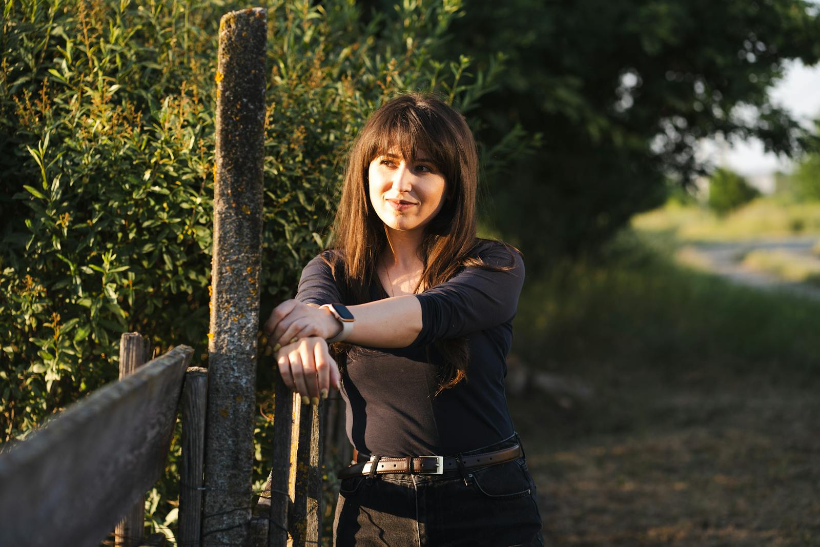 A young woman leans on a fence in the countryside during a beautiful sunset, enjoying the peaceful atmosphere.