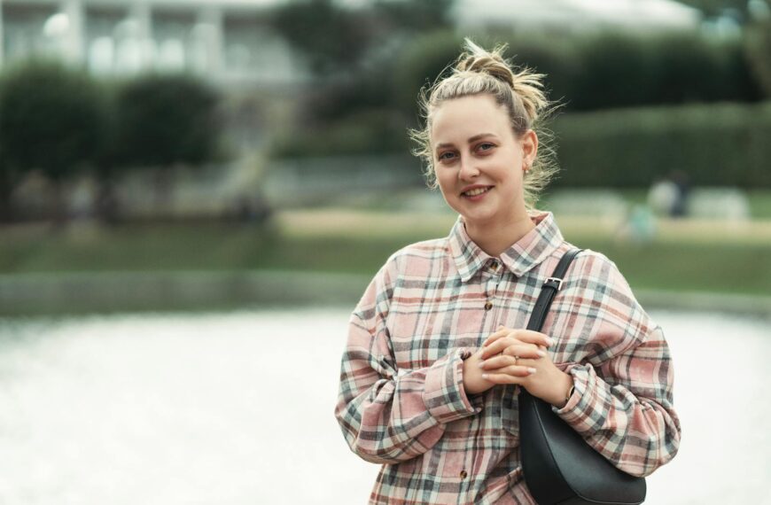 A woman in casual attire standing by a lake smiling outdoors, conveying a relaxed mood.