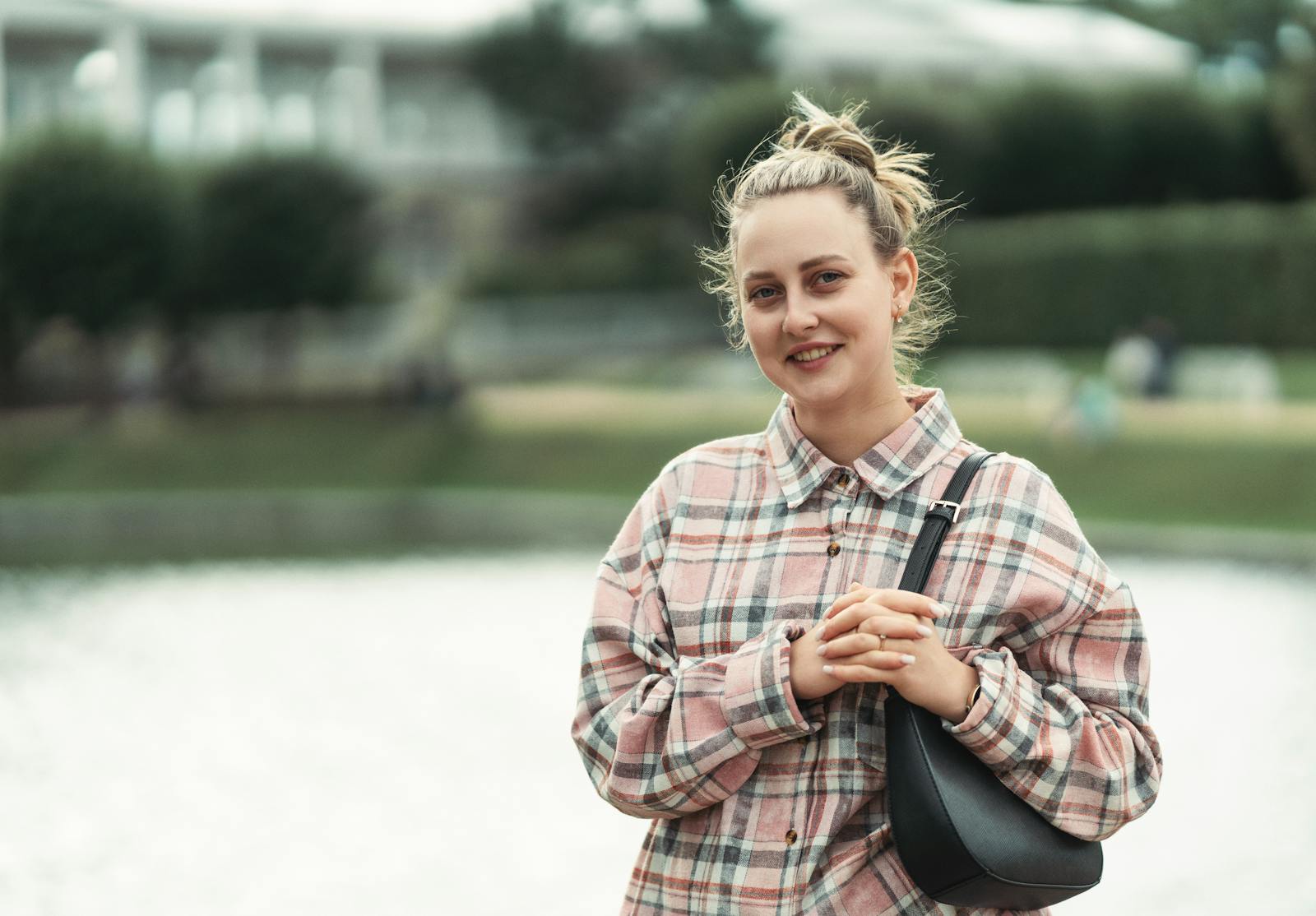 A woman in casual attire standing by a lake smiling outdoors, conveying a relaxed mood.