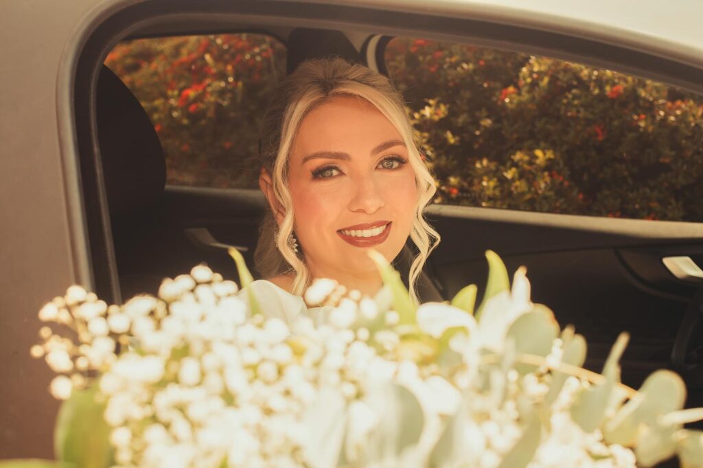 Smiling bride looking out of car window with bouquet in foreground, sunny day.