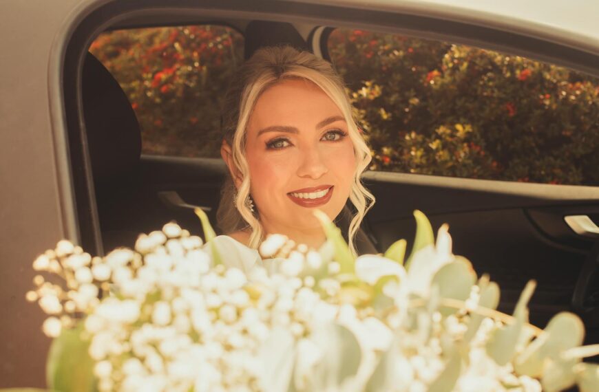Smiling bride looking out of car window with bouquet in foreground, sunny day.