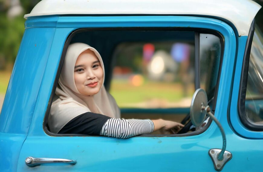 A smiling woman in a hijab sits in a classic blue truck, enjoying a sunny day.